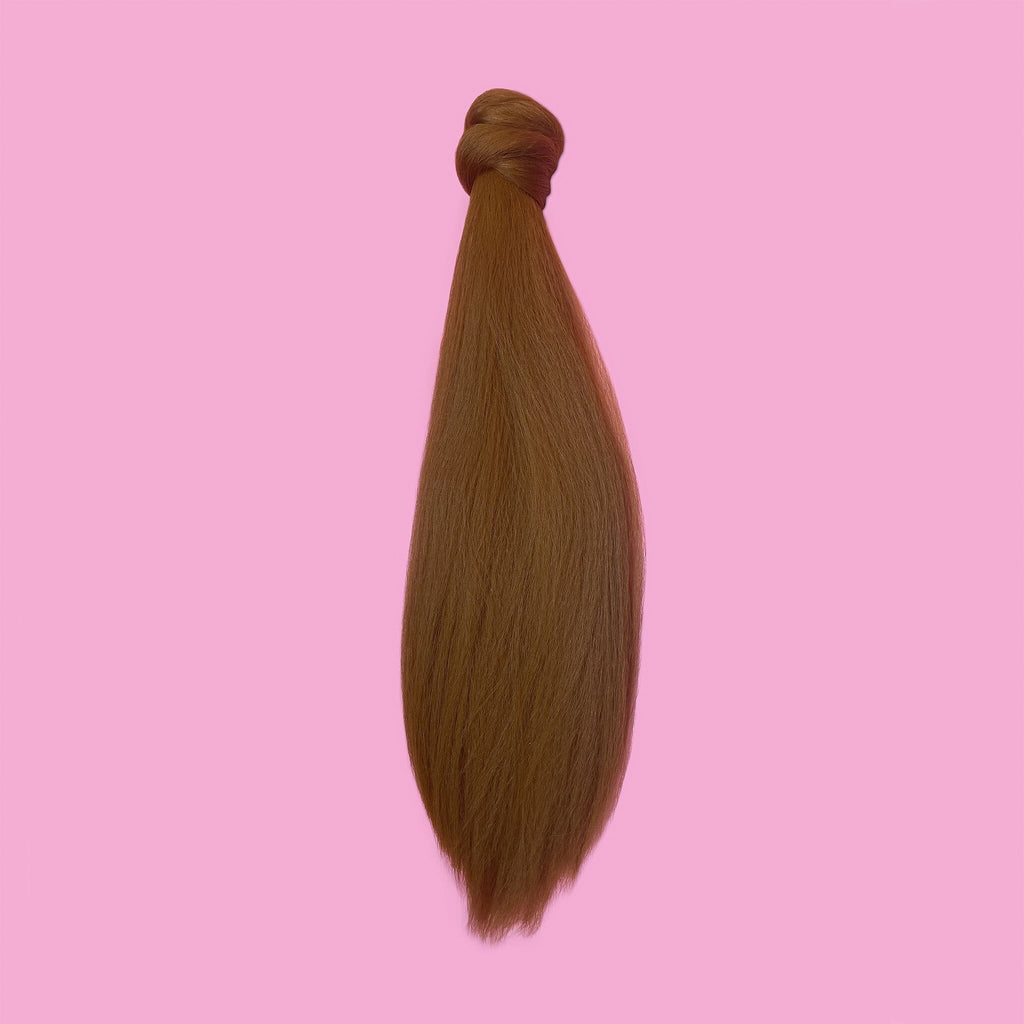 The Big Glam Nation Blowout Ponytail is laid flat with against a pink backdrop. It is "top knotted" and has a curvaceous flow down to the ponytail tip in a cumin-like brownish with yellow hue color. The photo shows the ponytails thick and kinkier texture - as if blow-dried.