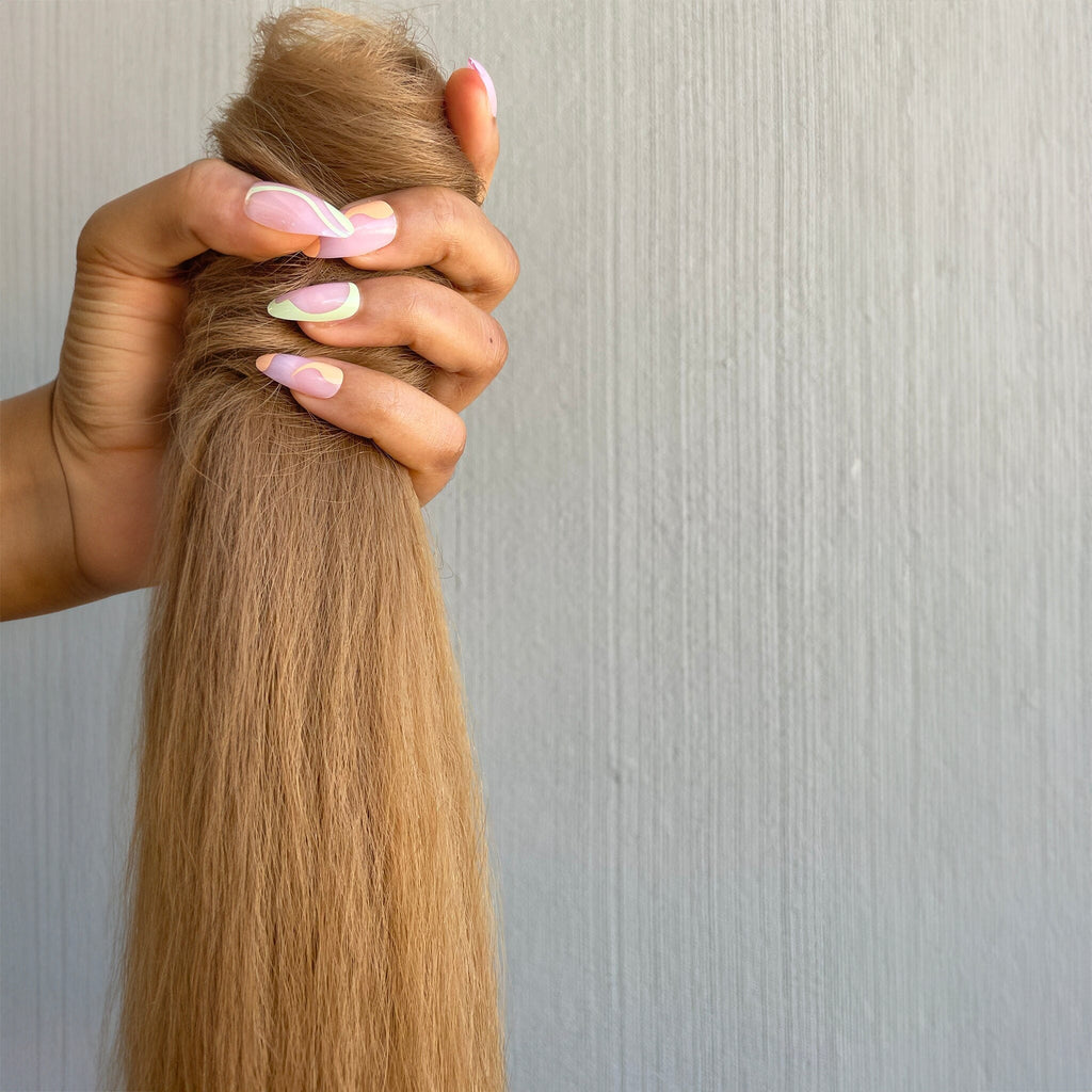 A model's hand holds the Big Glam Nation Blowout Ponytail in a close up photo against a grey wall. The photo emphasizes the thick and kinkier texture - as if blow-dried.