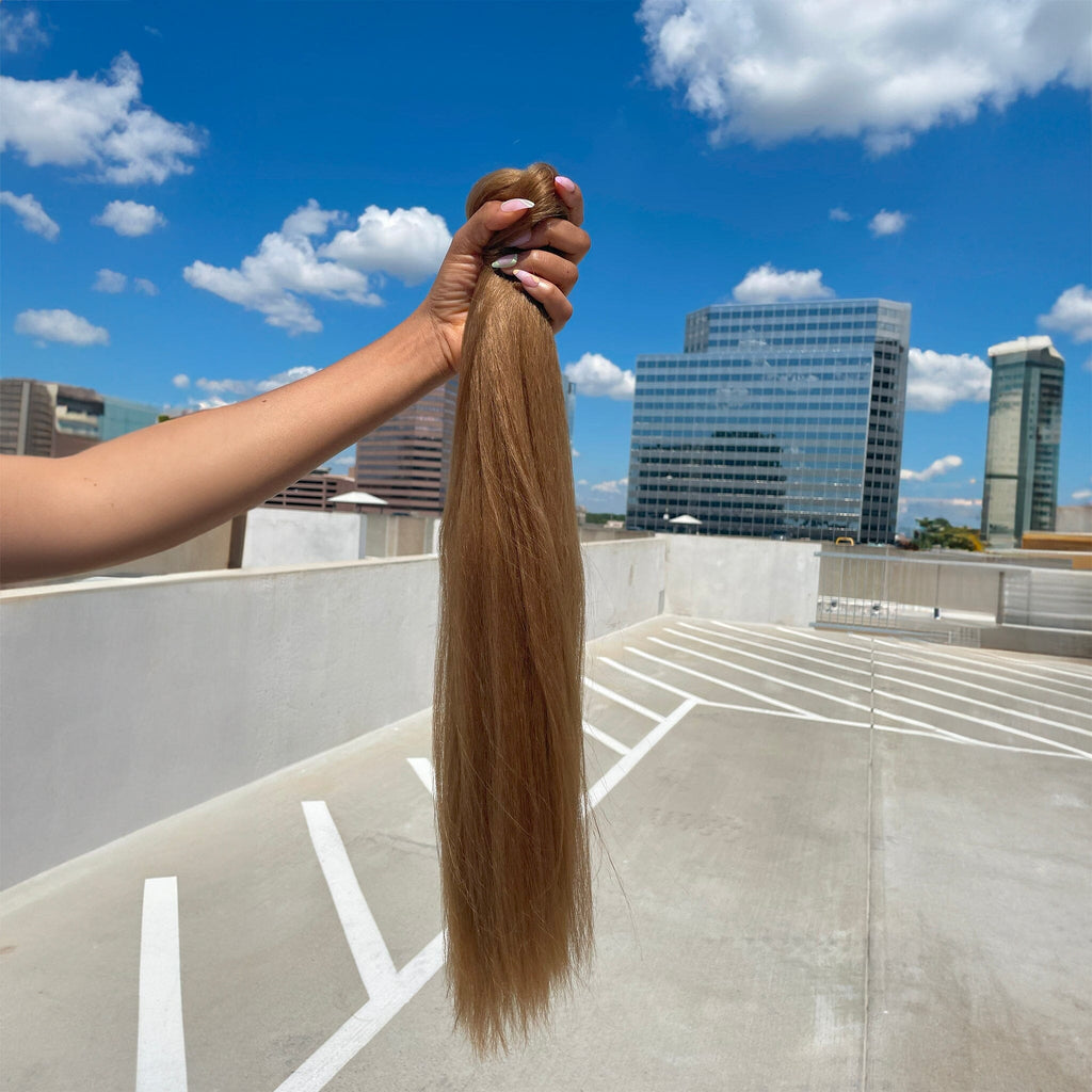 A model's hand holds the Big Glam Nation Blowout Ponytail against a sky background. The photo emphasizes the bold color, specifically - the ponytail's yellow-brown hue - in sunlight.