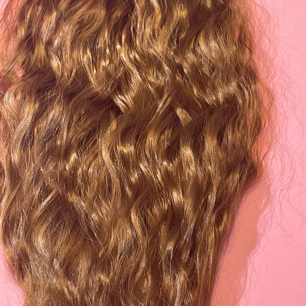 Ponytail Extensions in Big Glam Nation’s ginger-brown color with a wavy texture and velcro wraparound application are laid flat on a warm pink background.