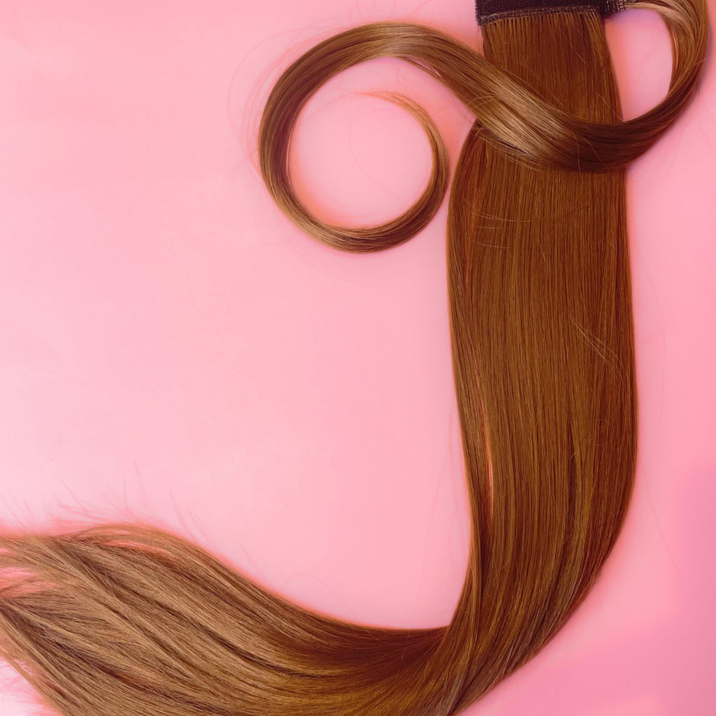 The Big Glam Nation Sleek Ponytail extensions are laid flat against a pink background in the color Ginger Brown (Deep Tint). Length and shine of the extensions are highlighted.