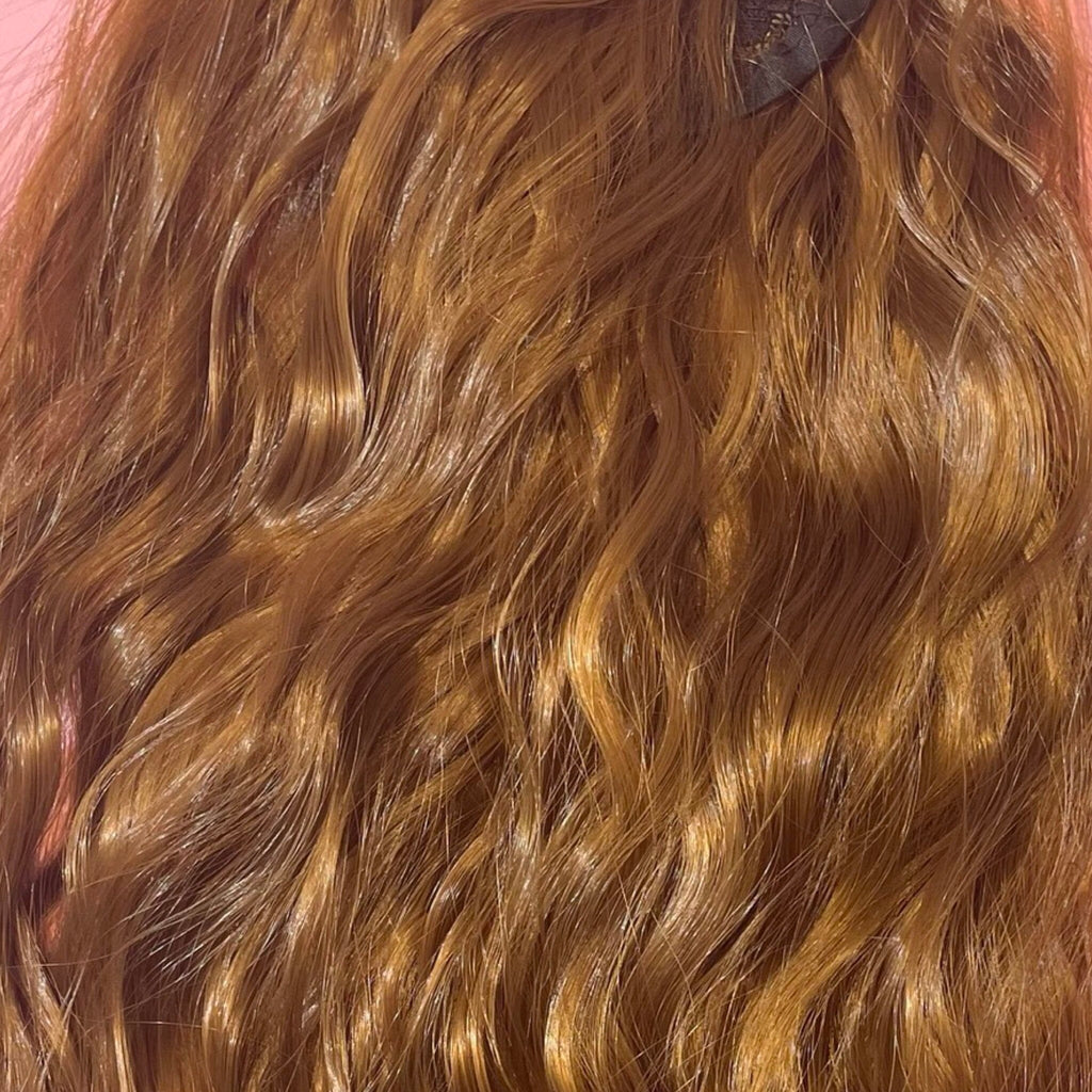 Ponytail Extensions in Big Glam Nation’s ginger-brown color with a wavy texture and velcro wraparound application are laid flat on a warm pink background.