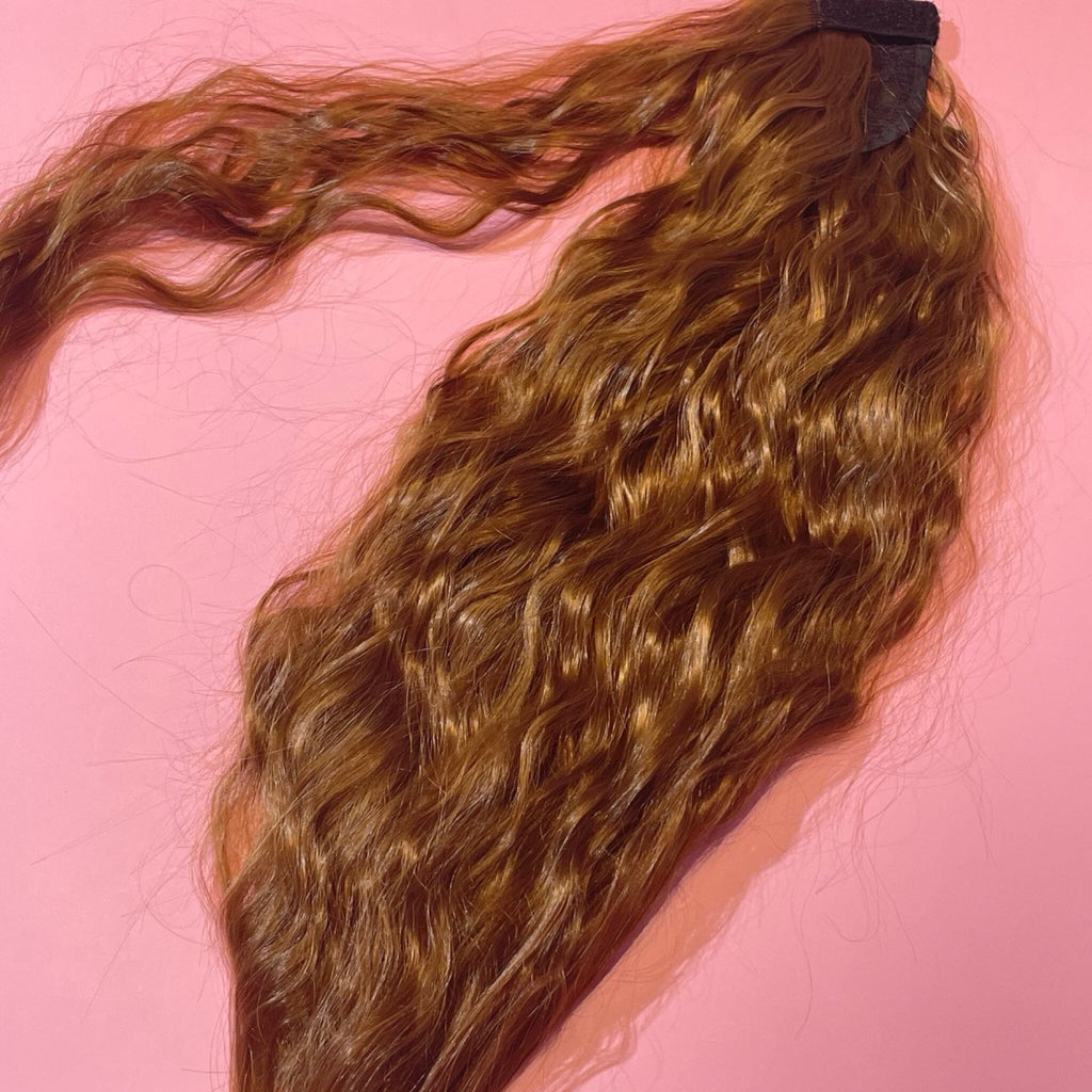 Ponytail Extensions in Big Glam Nation’s ginger-brown color with a wavy texture and velcro wraparound application are laid flat on a warm pink background.