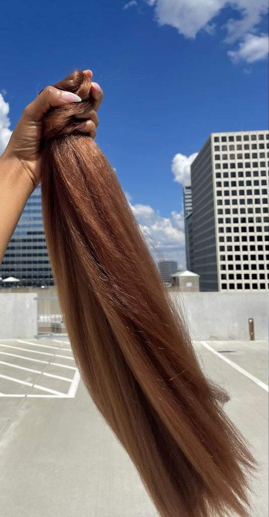  Big Glam Nation Blowout Ponytail extension held upagainst a sky background. The photo emphasizes the hair extensions' bold color, specifically - the ponytail extension's reddish orange hue - in sunlight.