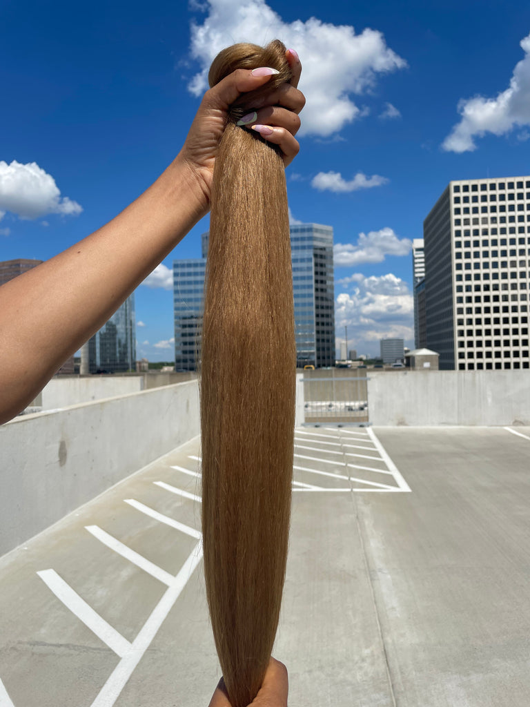 A model's hand holds the Big Glam Nation Blowout Ponytail against a sky background. The photo emphasizes the bold color, specifically - the ponytail's yellow-brown hue - in sunlight.