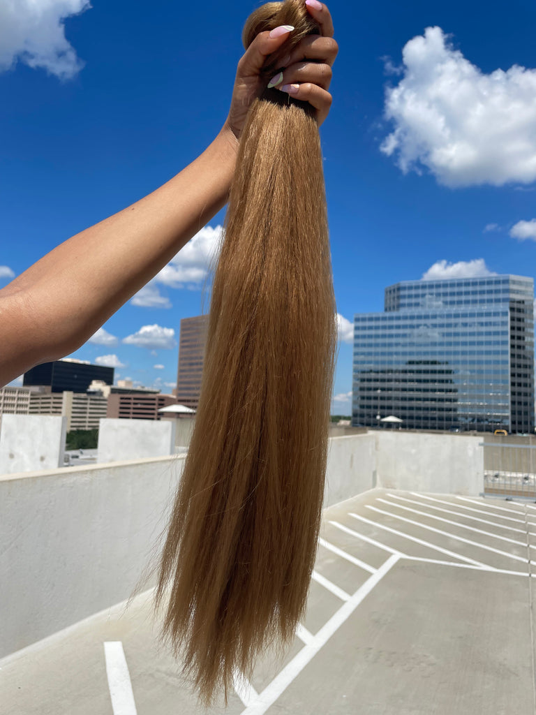 A model's hand holds the Big Glam Nation Blowout Ponytail against a sky background. The photo emphasizes the bold color, specifically - the ponytail's yellow-brown hue - in sunlight.